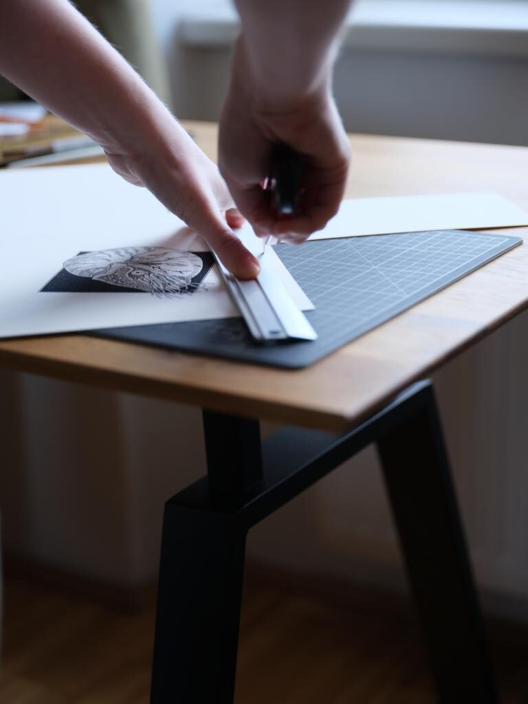 Es sind Hände zu sehen. Die eine hält ein Metalllineal, die andere ein Teppichmesser, mit dem gerade das Papier einer Tintenzeichnung auf einer Schneideunterlage zugeschnitten wird.