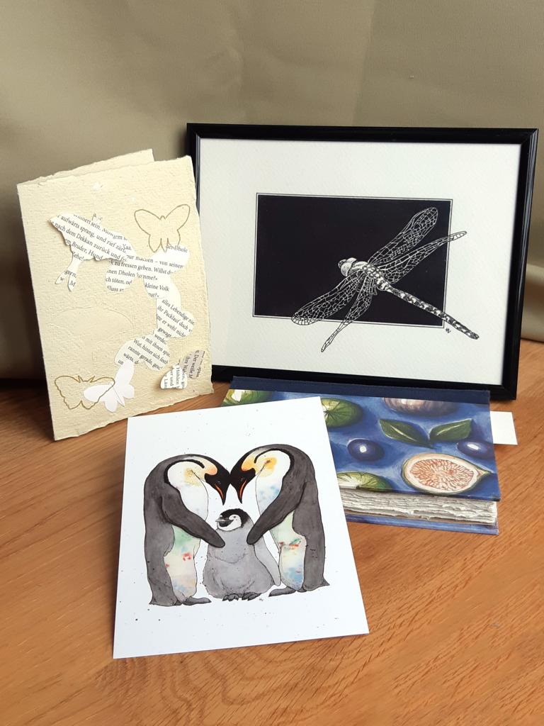 Zu sehen sind eine Grußkarte aus handgeschöpftem Papier mit Schmetterlingen, eine gerahmte Tintenzeichnung einer Libelle, ein Print eines Aquarells von Pinguinen und ein kleines Notizbuch, aus dem ein Lesezeichen herausschaut.