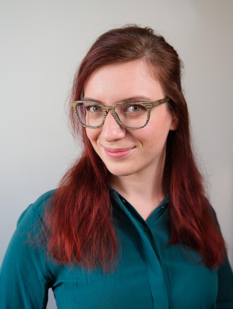 Portrait von Veronika Hantschel alias tinteronika. Sie trägt eine türkise Bluse, eine hölzerne Brille, hat rote Haare und lächelt verschmitzt.