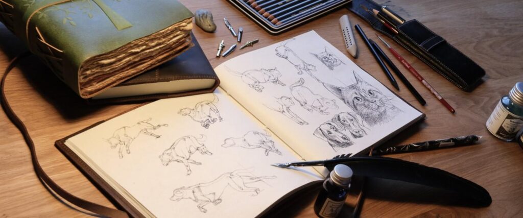 Es ist eine Schreibtischfläche zu sehen, auf der Kunstutensilien liegen: Skizzenbücher, eines davon aufgeschlagen mit Skizzen von Hunden und Katzen, Zeichenfedern, ein Tintenfass, Stifte