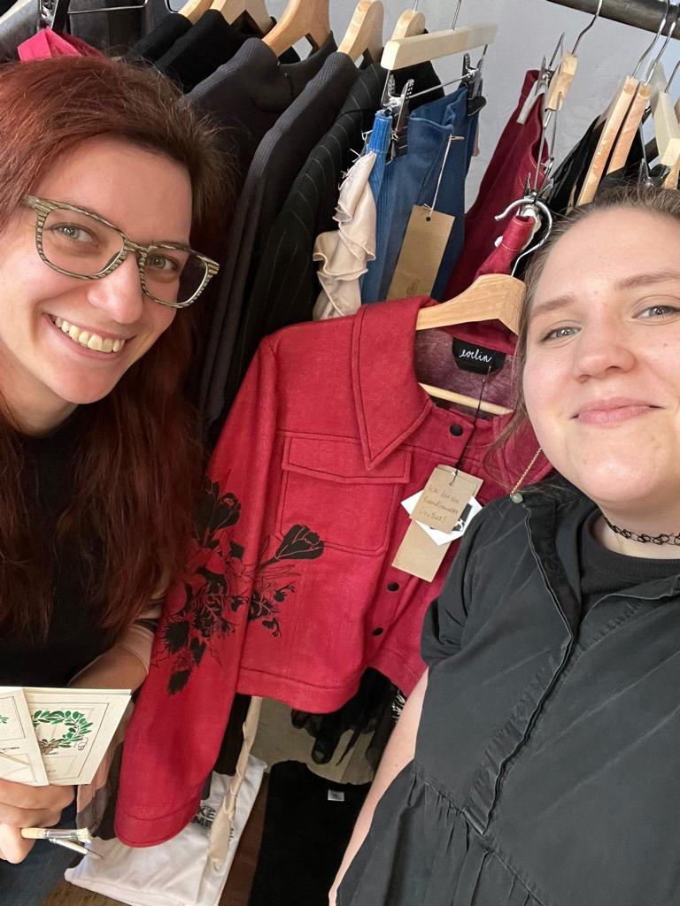 Die Illustratorin Veronika Hantschel und die Designerin Evelin Grubbauer lächeln in die Kamera. Hinter ihnen ist eine Kleiderstange mit Gewand zu sehen, eine rote Jacke mit der Bemalung eines schwarzen Blumenmotivs steht dabei im Mittelpunkt.