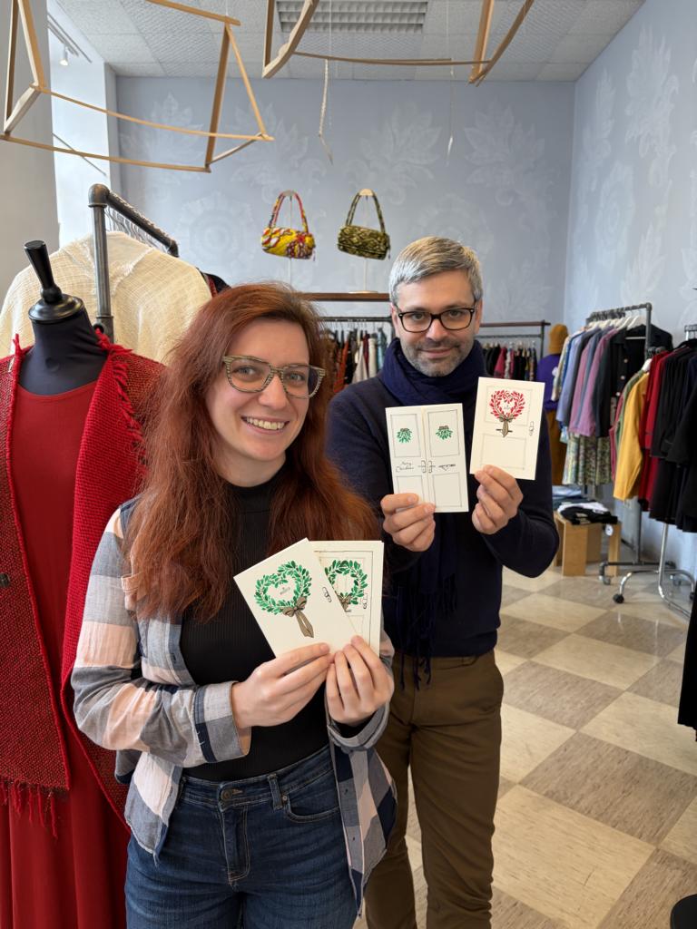 Die Illustratorin Veronika Hantschel und der Designer Gabriel Baradee halten Grußkarten hoch, auf denen Türkränze zu sehen sind. Im Hintergrund ist der Shakkei Design Store und Mode zu sehen.